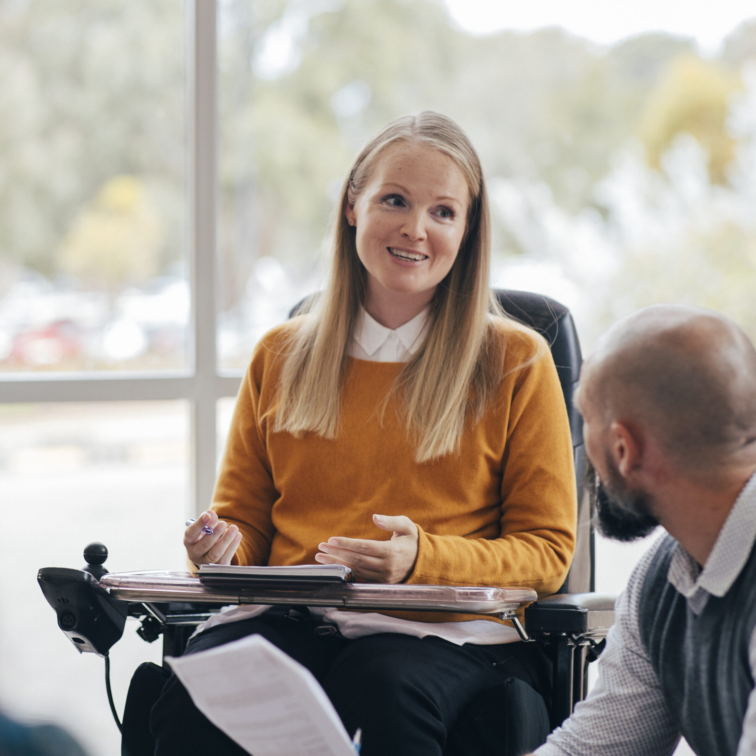 A woman looks happy and confident as she leads a group discussion at her place of work. She is a wheelchair user and has Muscular Dystrophy.